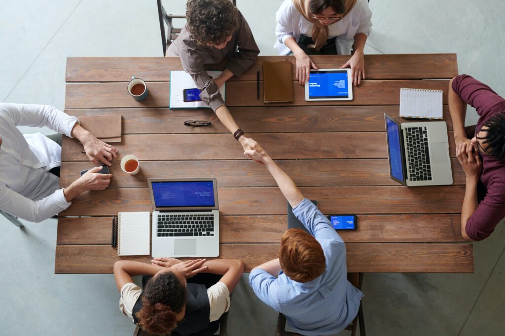 People with laptops sitting at table, two of them shaking hands across the table.