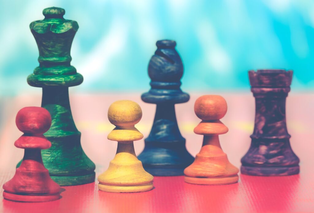 Six wooden chess pieces on a red table with a blue blurred background; a green king, blue bishop, purple rook, and red, yellow, and orange pawns.